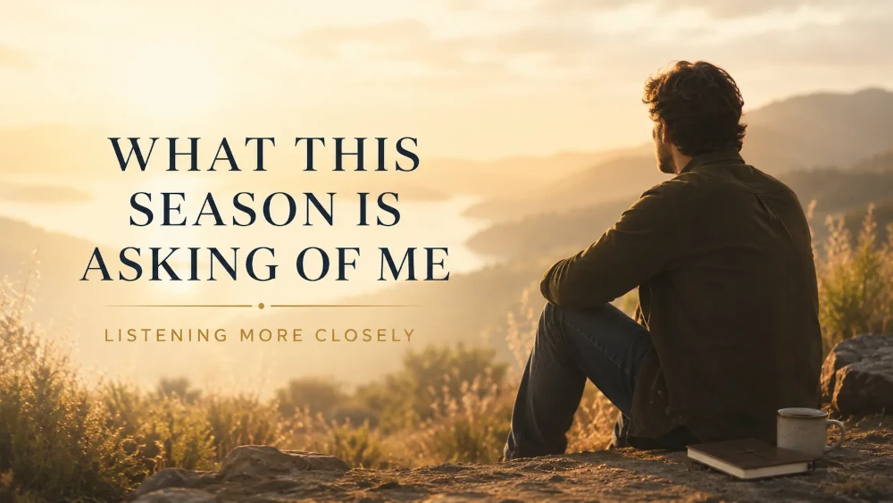 A man sits on a hillside at golden hour, looking over a quiet lake and mountains with a journal and mug beside him.